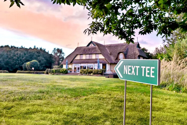 Exterior view of the golf center at sunset. In the foreground, a sign points to the next tee.