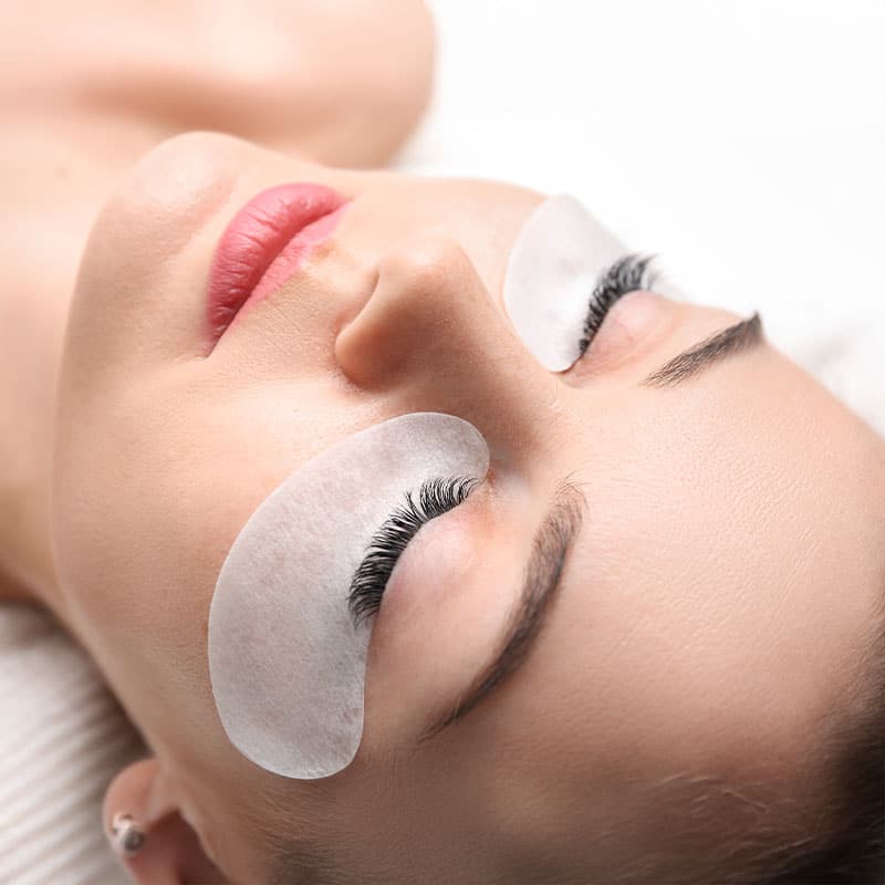 A woman relaxes while her eyelash care works on protective pads.