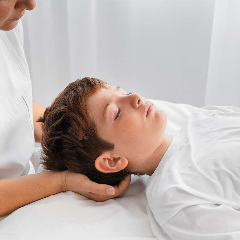 A teenager lies with closed eyes on a treatment table at Arkona Strandhotel Rügen, while a therapist gently holds his head.