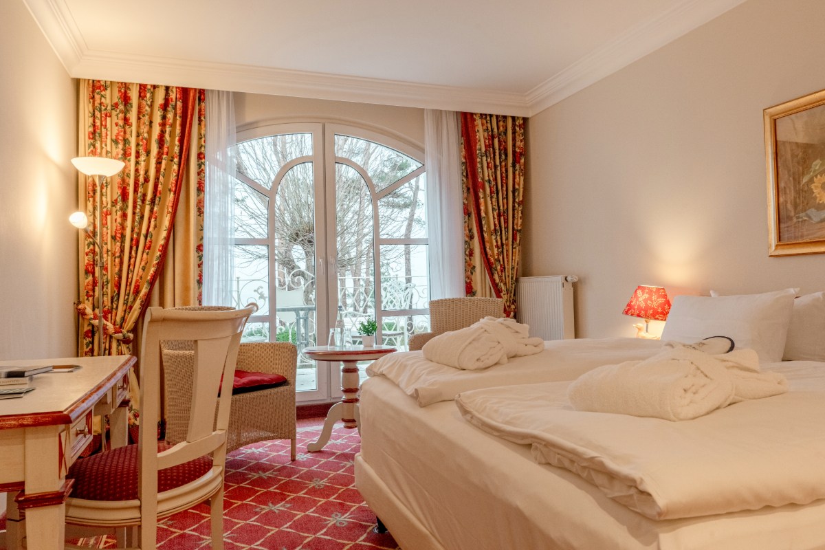 Großes Zimmer im Hotel mit Doppelbett, Schreibtisch und Sitzmöbeln und einem großen Fenster mit Balkon.