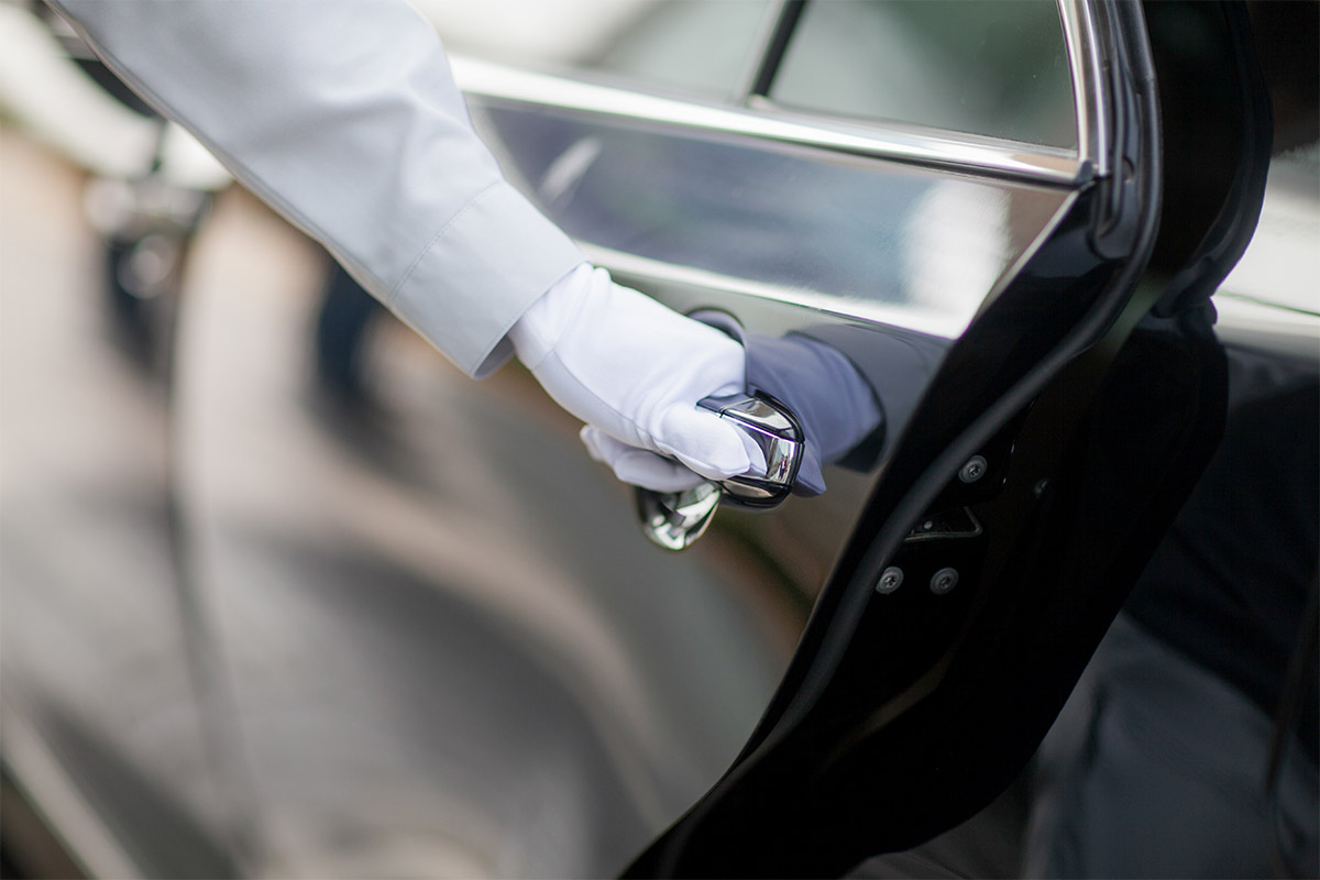 Nahaufnahme der behandschuhten Hand eines Hotelangestellten beim Öffnen einer dunklen Autotür.